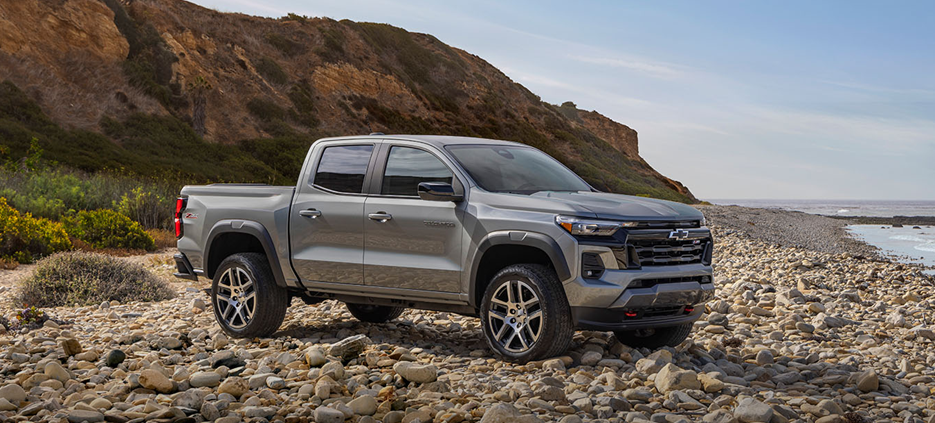 2025 Chevrolet Colorado midsize pickup truck in gray parked on a rocky coastal shoreline with cliffs and ocean in the background.