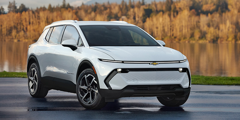 A sleek white Equinox-EV is parked on a wet surface with autumn trees reflecting in a lake behind it. The vehicle exudes a modern and robust design.