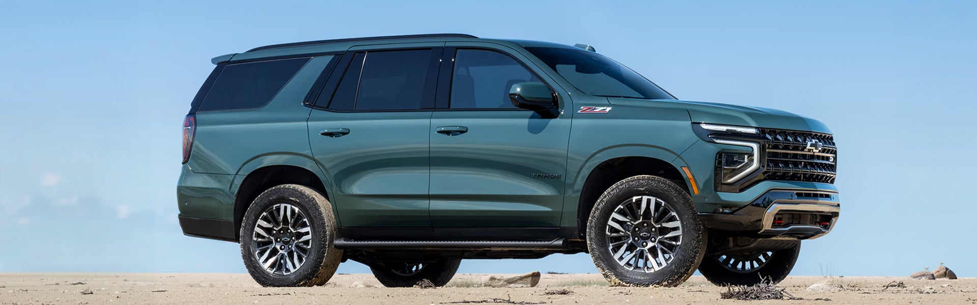 2026 Chevy Tahoe Z71 SUV shown in a side-front view, featuring a bold grille, rugged tires, and a dark green exterior against an open landscape.